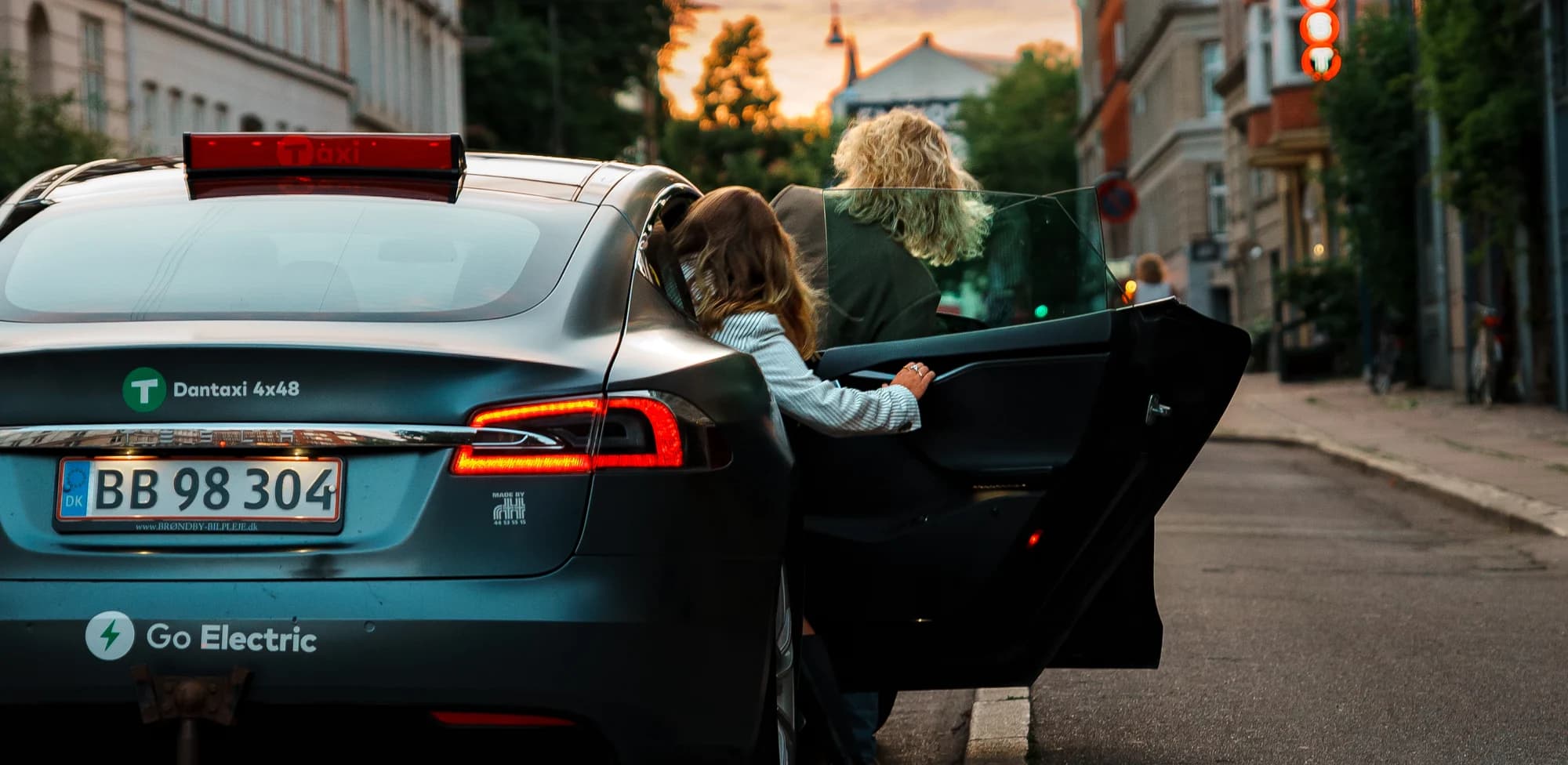 A passenger opening the door of an electric Dantaxi at sunset.
